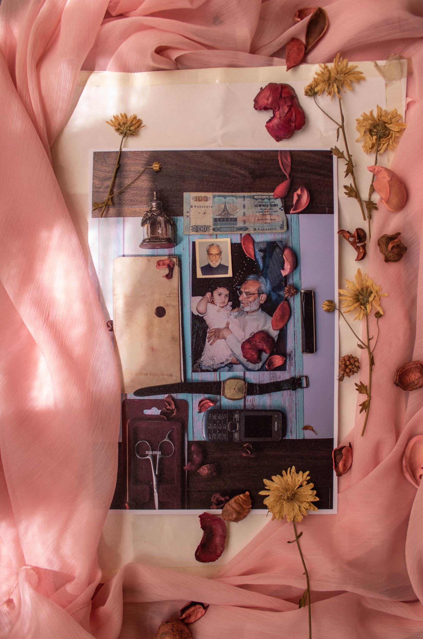 A printed collage of a grandfather holding a baby, surrounded by a banknote, a wristwatch, a keypad phone and scissors, taped onto pink chiffon and strewn with dried petals