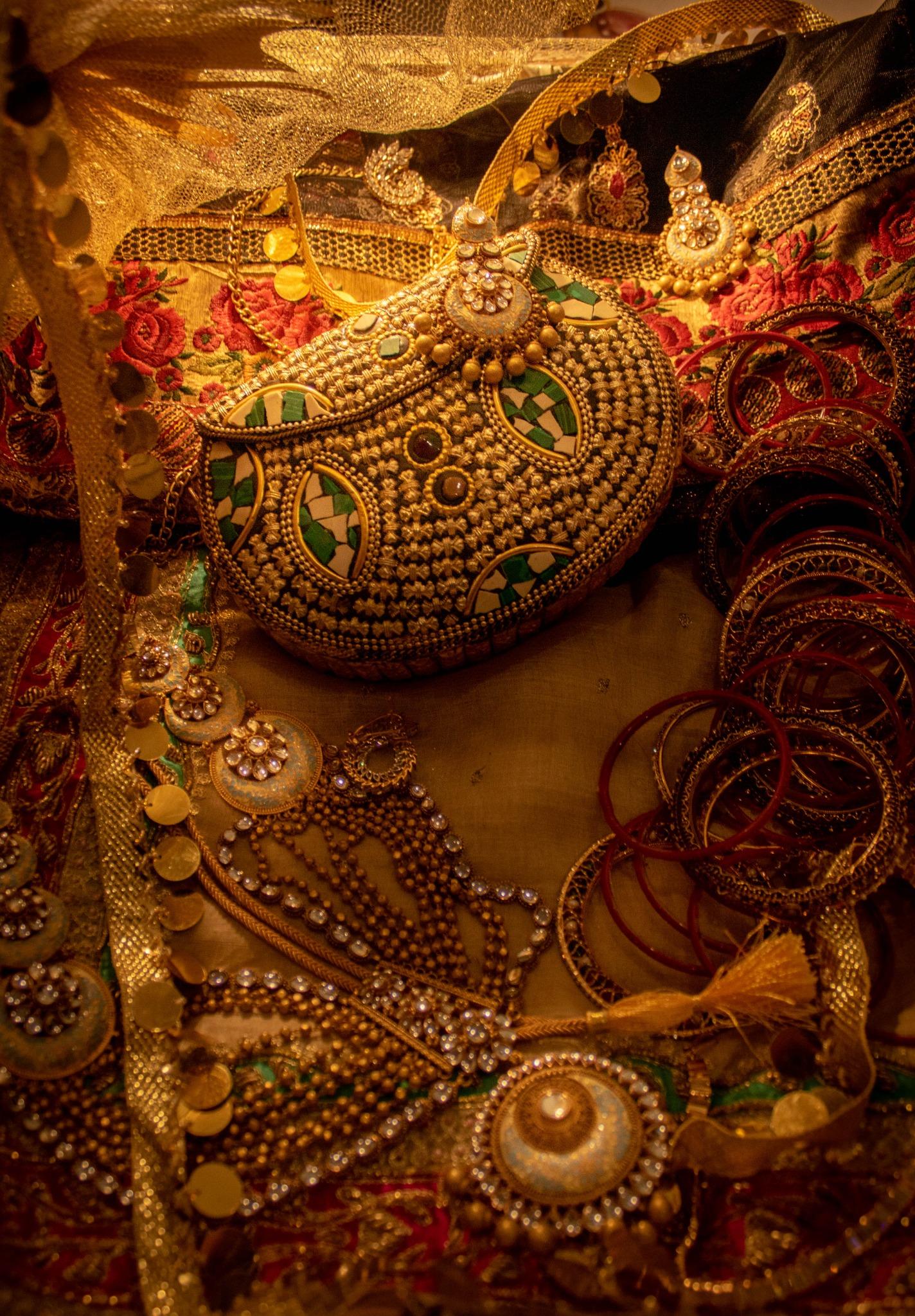 Warm lamplight on gold bridal jewellery, kundan earrings, a beaded purse and stacks of red and gold bangles heaped on embroidered cloth