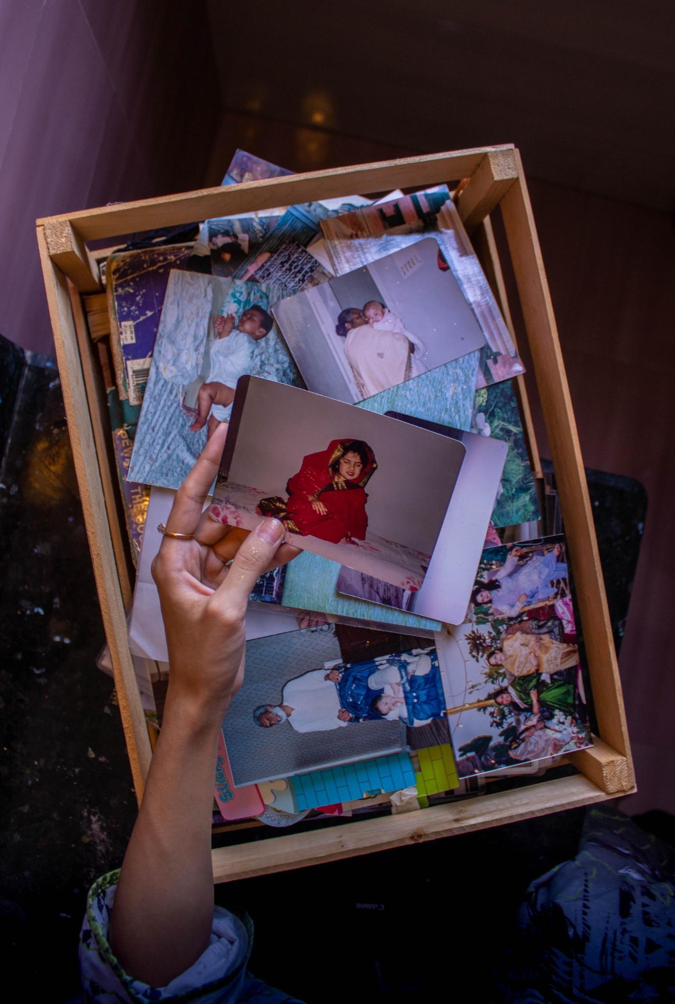 A hand holding up a wooden tray filled with old family photographs, one print of a girl in a red dupatta pulled forward from the stack