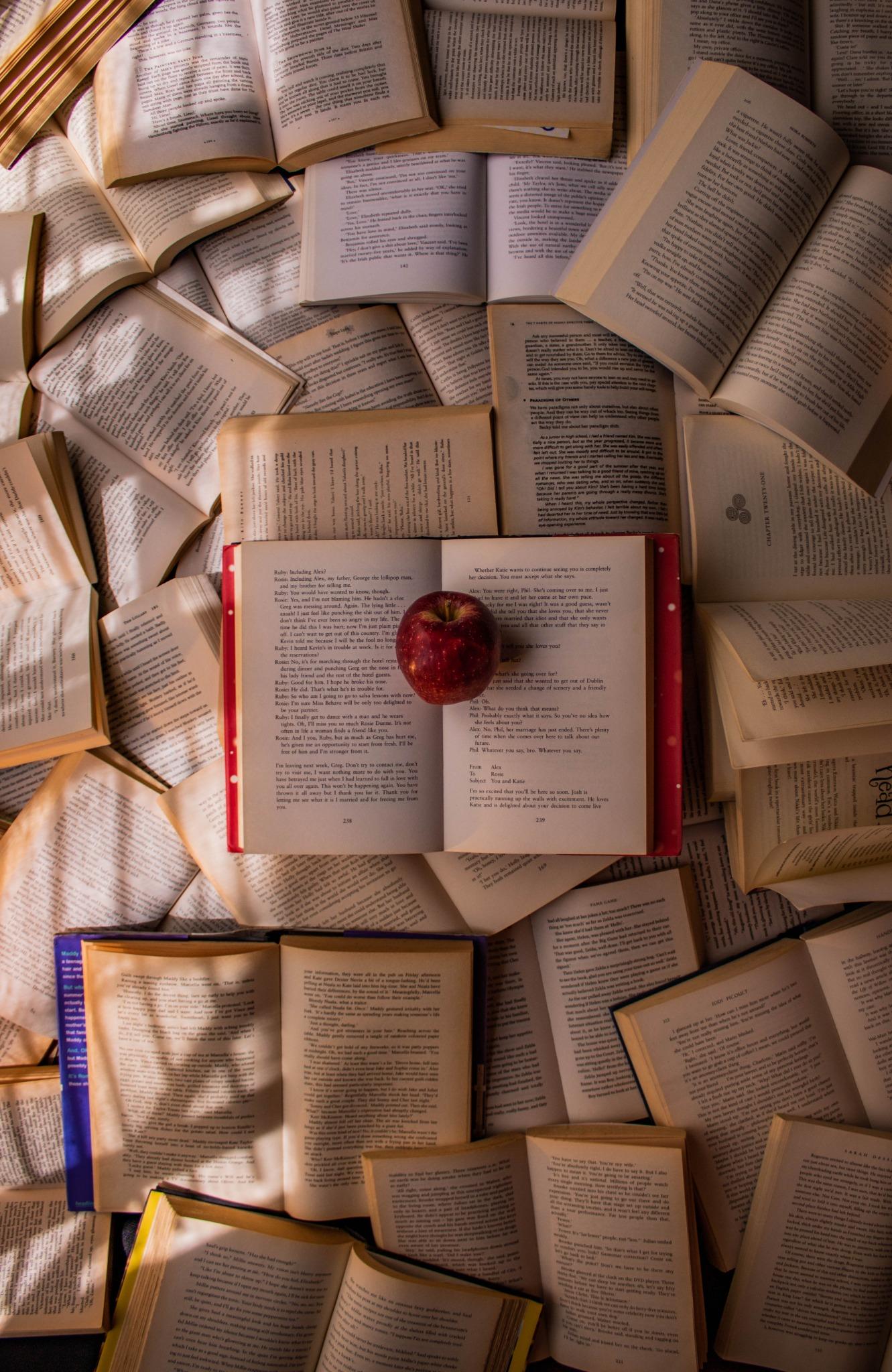 Dozens of open books laid face-up in a dense field, with a single red apple resting on the pages of the book at the centre