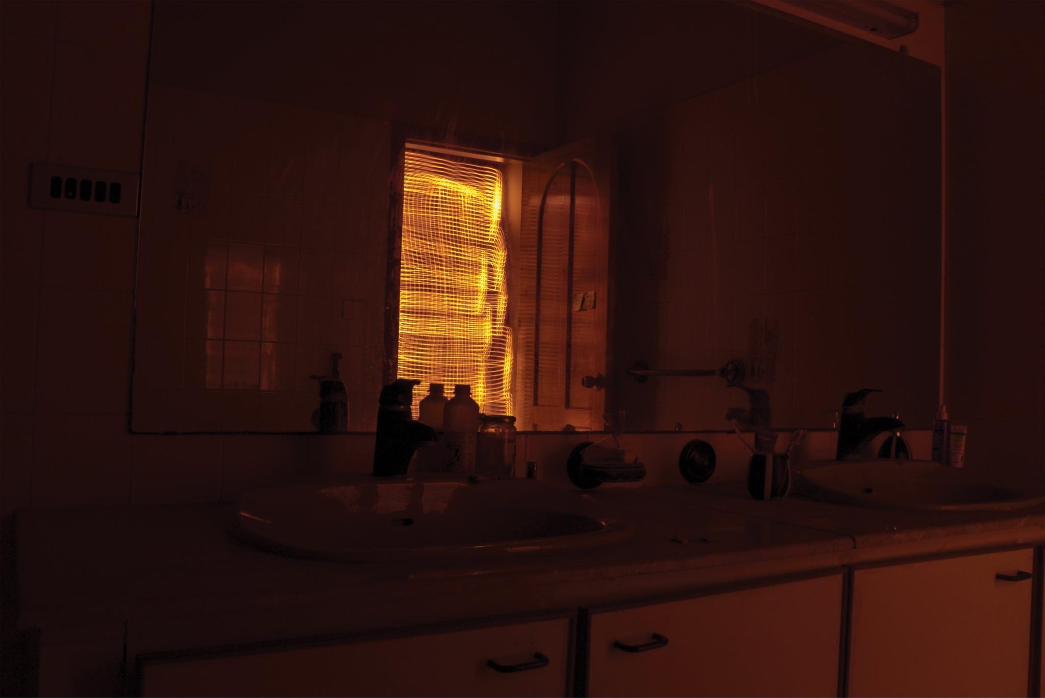 A dark bathroom mirror and basin with amber light striping through the shuttered window