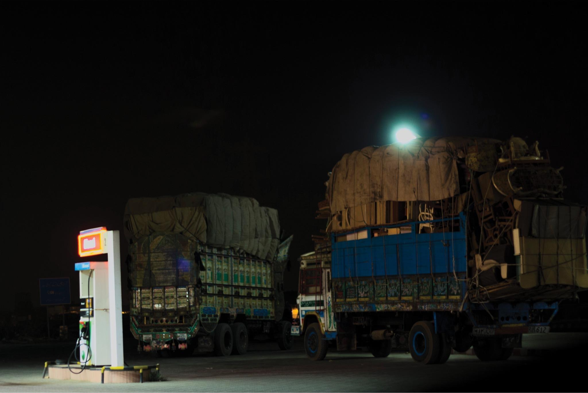 Two loaded cargo trucks parked at a lit petrol pump under a full moon at night