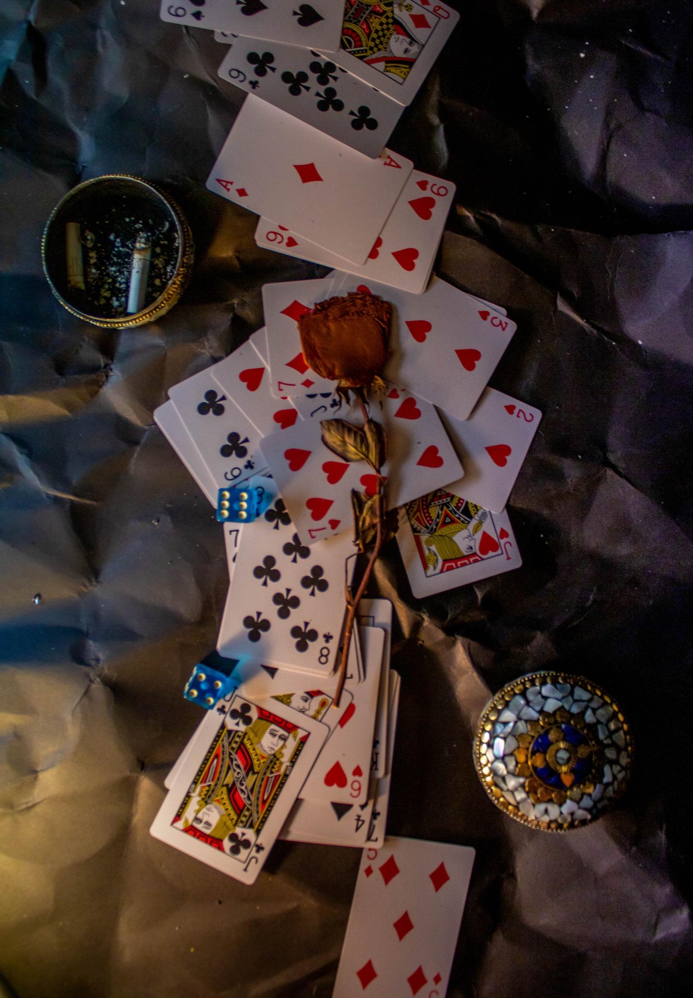 Playing cards scattered over crumpled dark paper with a dried rose across them, two blue dice, a mosaic lid and an ashtray holding two cigarette ends