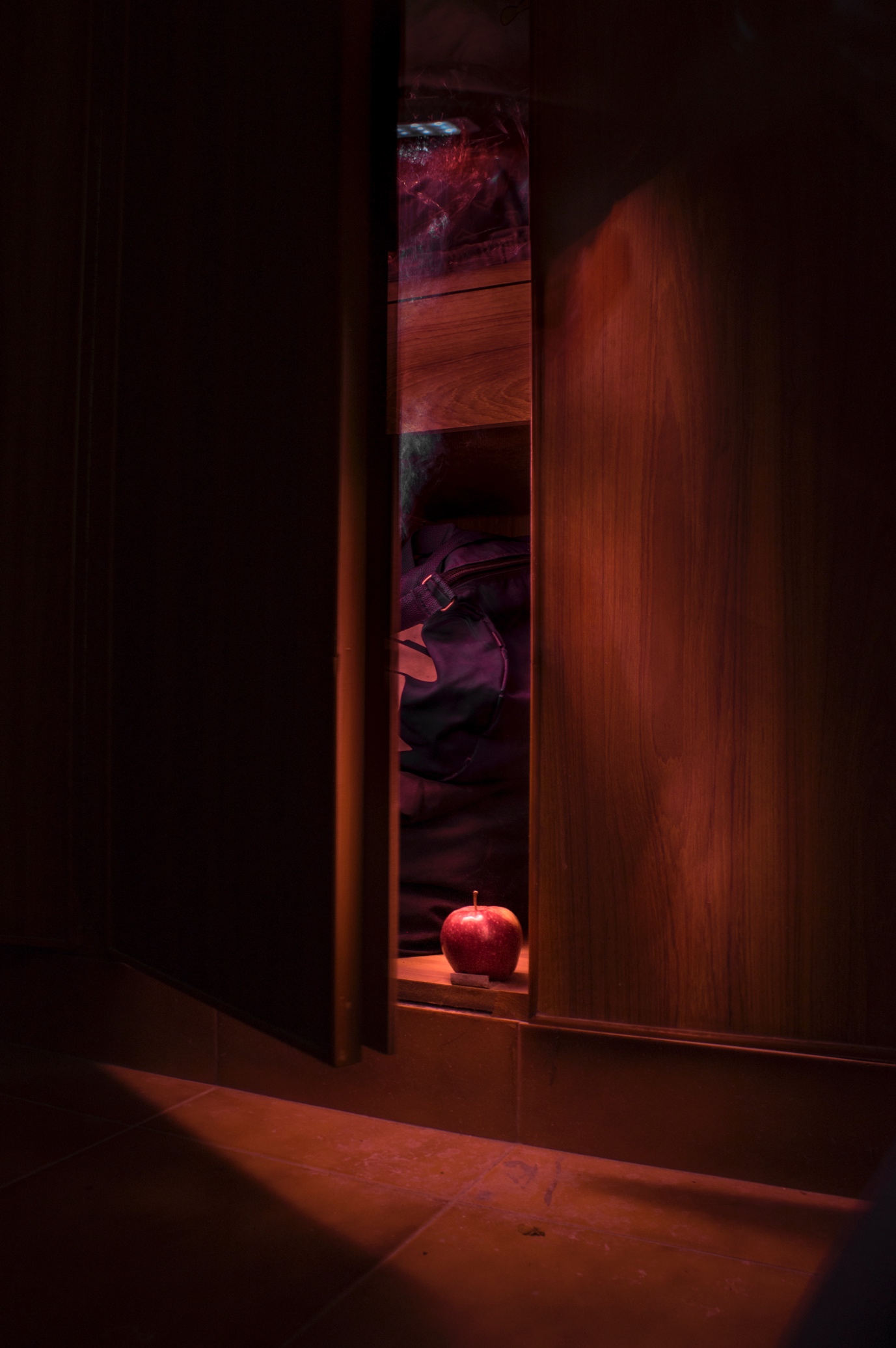 A red apple on the ledge of a half-open wooden cupboard, lit red from inside, clothes hanging behind it