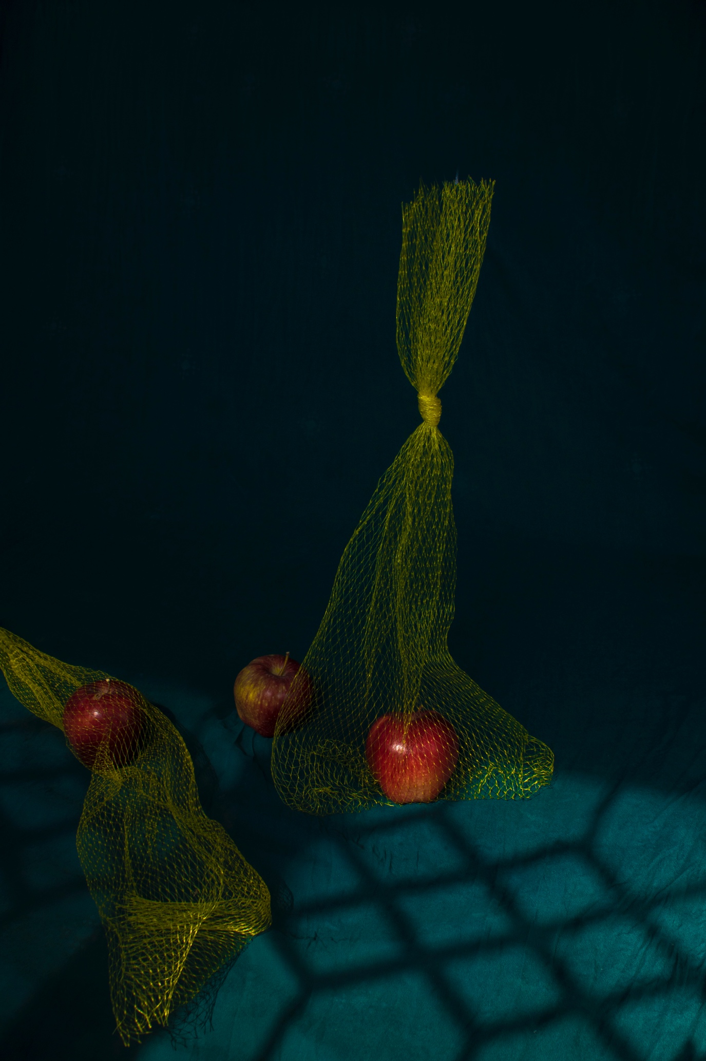 Three red apples in yellow mesh market bags on dark teal cloth, one bag hanging, a barred beneath