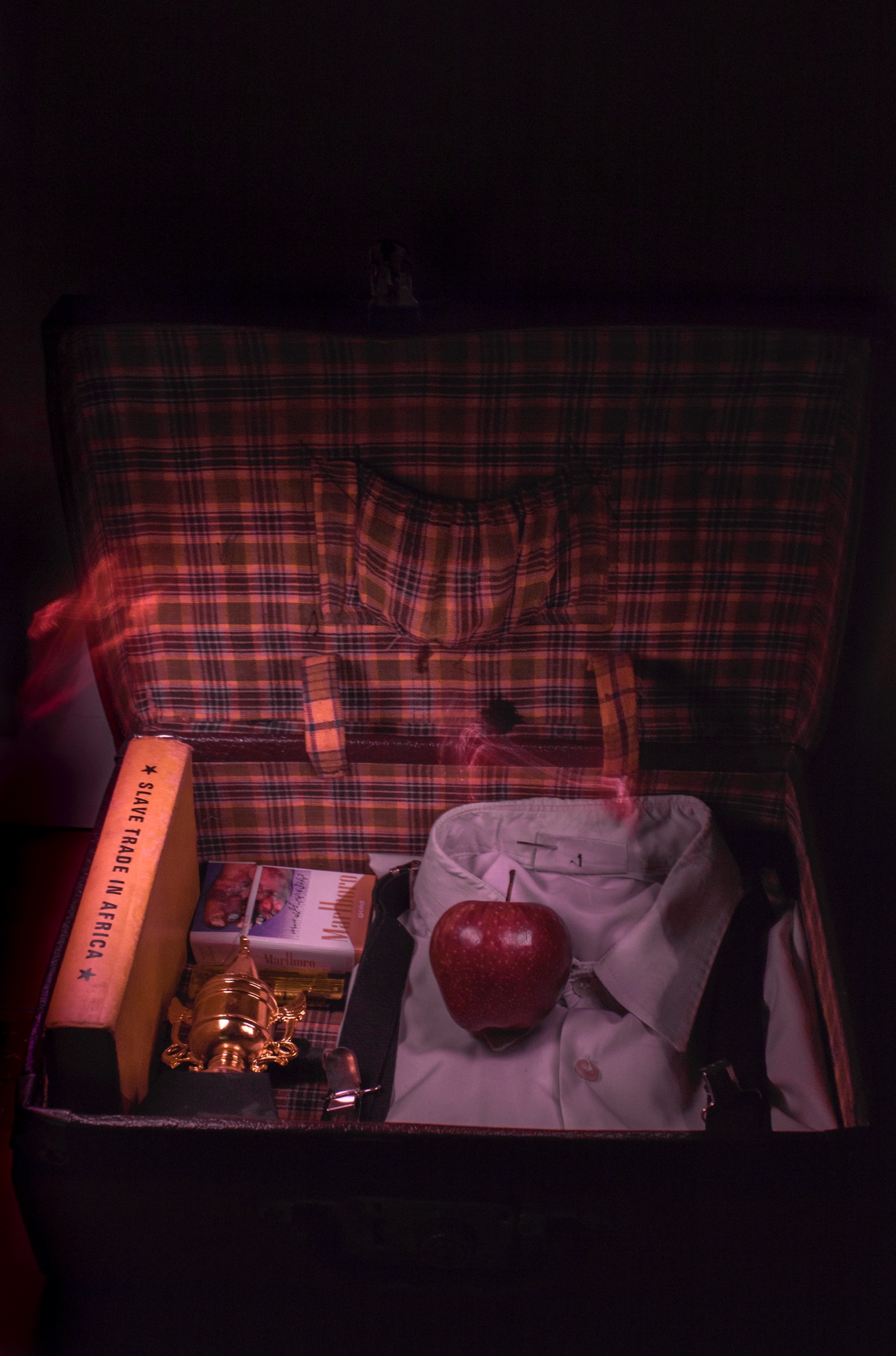 An open tartan-lined travel trunk holding a red apple, a folded white shirt, a brass trophy and a book titled Slave Trade in Africa